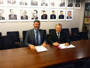 Dwóch mężczyzn siedzi za podłużnym stołem, podpisując dokumenty.