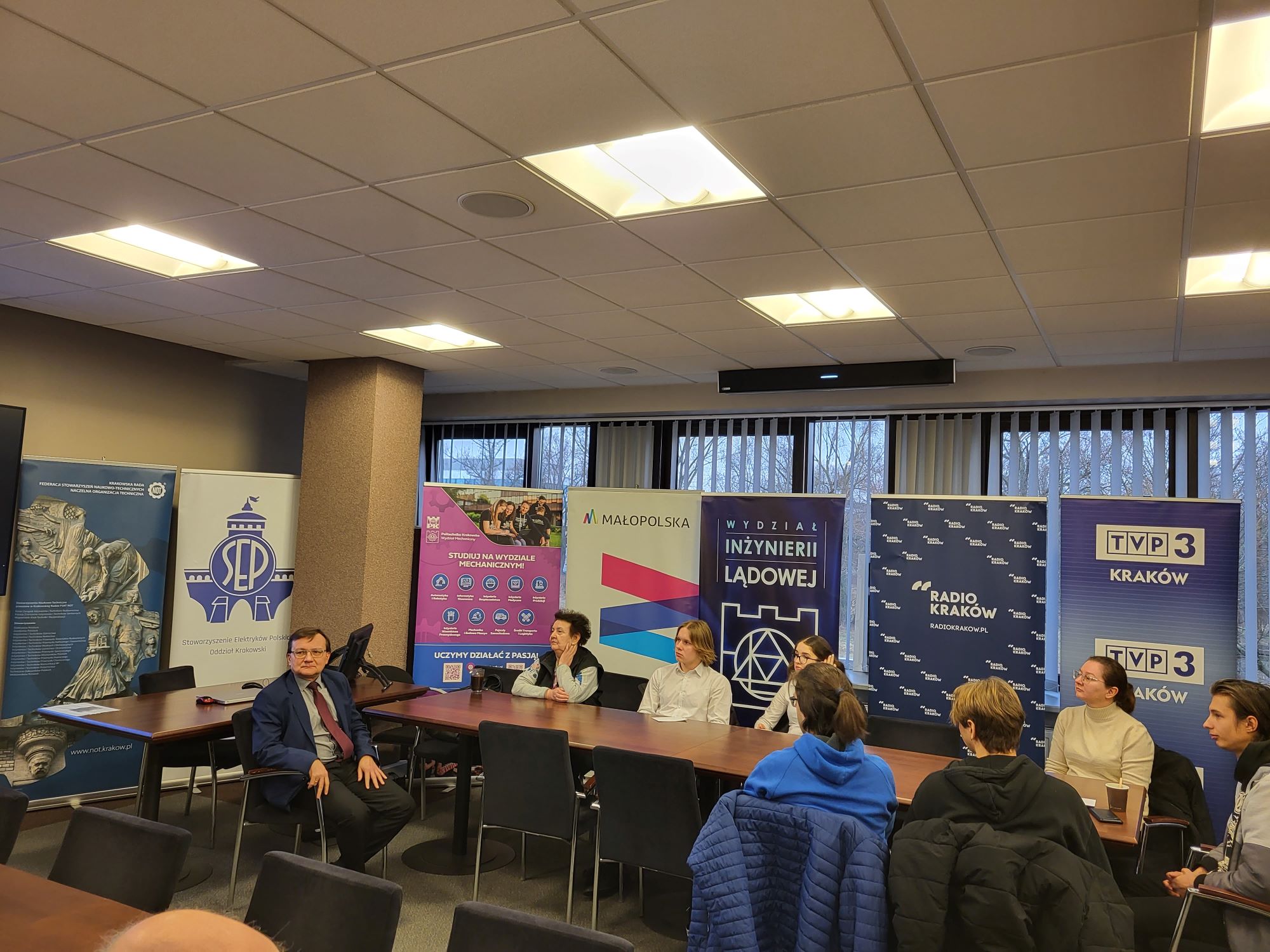 Młodzi ludzie siedzą przy stołach. W tle rollupy różnych instytucji 0 Radio Kraków, TVP3, Województwa Małopolskiego, Wydziału Inżynierii Lądowej Pk i Wydziału Mechanicznego PK