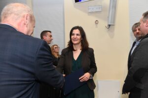 Kobieta w brązowej marynarce odbiera dyplom z rąk mężczyzny, stojącego tyłem do aparatu.