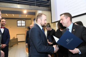 Na pierwszym planie dwóch mężczyzn wymienia uścisk dłoni. Są elegancko ubrani. Na drugim planie więcej osób.