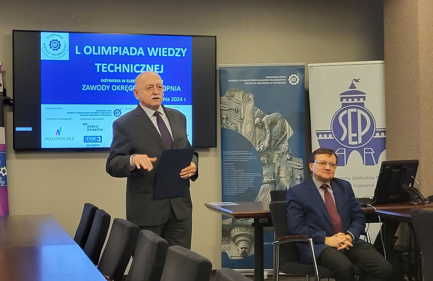 Dwóch mężczyzn, jeden stoi i przemawia, drugi siedzi na krzesle. W tle Telebim z napisem L Wiedzy Technicznej, oraz dwa rollupy - NOT i SEP.