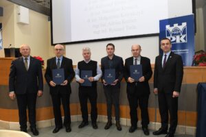 6 mężczyzn pozuje do zdjęcia, 4 z nich trzyma w rękach dyplomy. W tle telebim z wyświetlonymi nazwiskami.