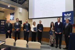 8 osób na zdjęciu, 6 z nich ma w ręku dyplomy, z tyłu telebim z wyświetlonymi nazwiskami