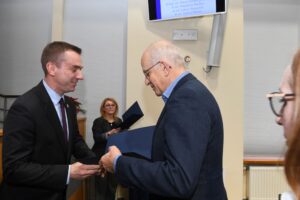 Dwóch, elegancko ubranych mężczyzn, jeden z nich wręcza drugiemu dyplom.