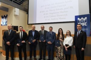 8 uśmiechniętych osób pozuje do zdjęcia, trzymają w rękach dyplomy. W tle telebim, z lewej strony roll-up z logiem PK.