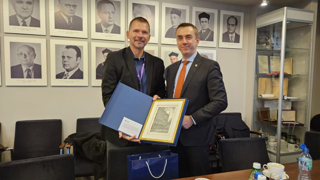 Dwóch mężczyzn ściska sobie ręce i wspólnie trzymają grafikę w okładce. Na grafice widać szkic budynku.