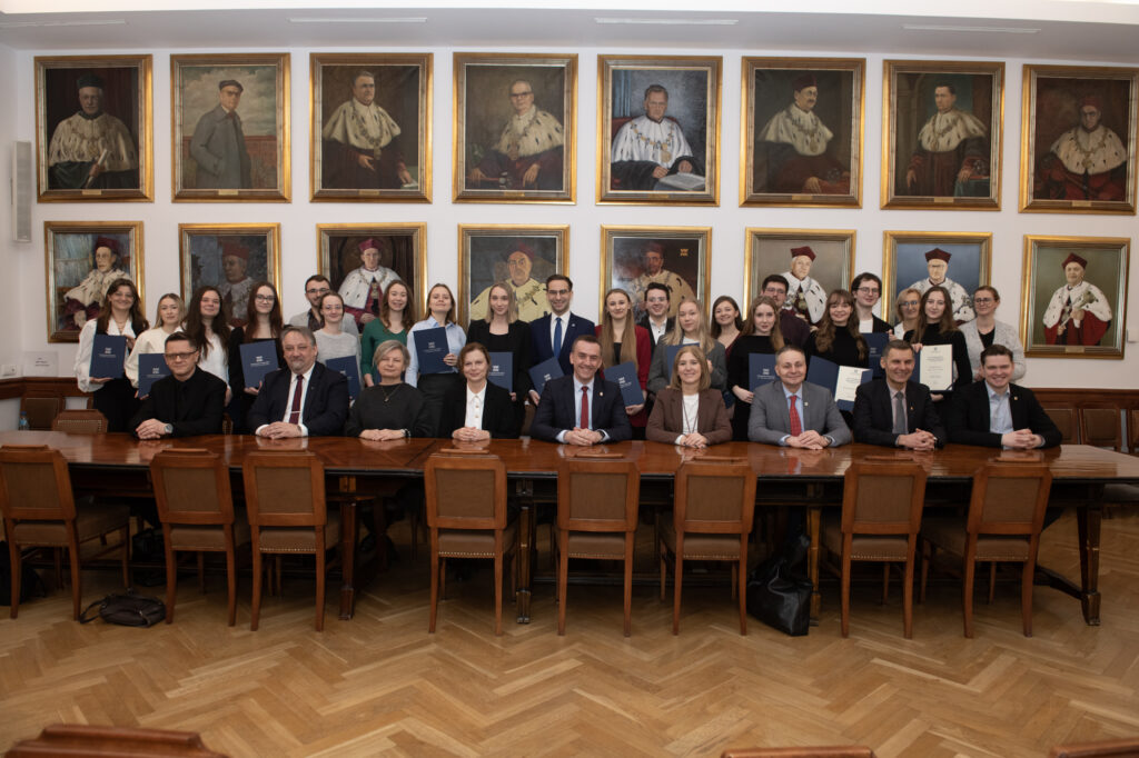 Grupa osób w różnym wieku i różnej płci pozuje do zdjęcia. Część osób siedzi za długim stołem, część osób stoi za nimi na tle ściany z portretami.