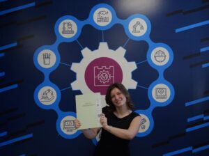 uśmiechnięta dziewczyna pozuje do zdjęcia ze swoją pracą inżynierską na tle grafiki z logo Wydziału Mechanicznego Politechniki Krakowskiej