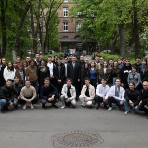 Grupa studentów pozuje do zdjęcia na dziedzińcu, przed popiersiem Tadeusza Kościuszki. W centralnej części zdjęcia uśmiechnięty mężczyzna. W tle zielone drzewa i budynek z cegły.