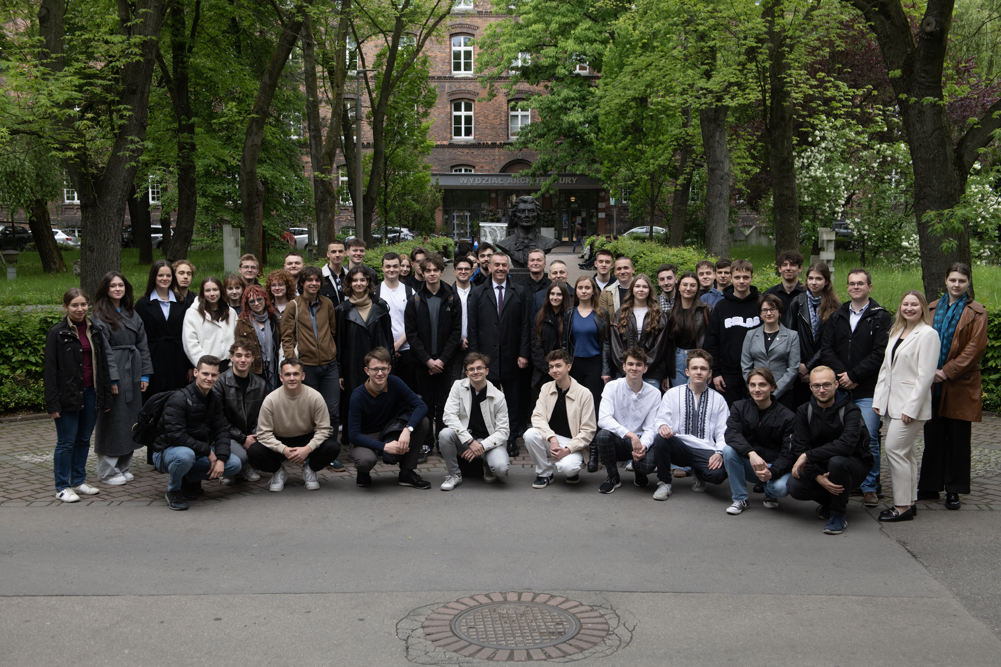 Grupa studentów pozuje do zdjęcia na dziedzińcu, przed popiersiem Tadeusza Kościuszki. W centralnej części zdjęcia uśmiechnięty mężczyzna. W tle zielone drzewa i budynek z cegły.