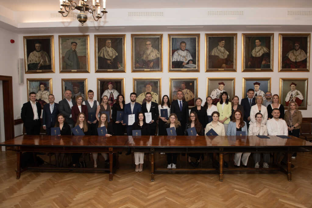 Grupa osób pozuje do zdjęcia na tle ściany z portretami. Przed nimi długi lakierowany stół. Część osób trzyma w rękach dyplomy w granatowych okładkach z logiem Politechniki Krakowskiej.