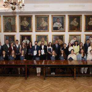 Grupa osób pozuje do zdjęcia na tle ściany z portretami. Przed nimi długi lakierowany stół. Część osób trzyma w rękach dyplomy w granatowych okładkach z logiem Politechniki Krakowskiej.