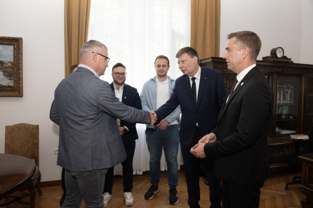 6 elegancko ubranych mężczyzn stoi w półokręgu. Dwóch z nich ściska sobie rękę. W tle brązowe meble w starym zabytkowym stylu.