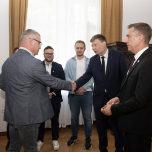 6 elegancko ubranych mężczyzn stoi w półokręgu. Dwóch z nich ściska sobie rękę. W tle brązowe meble w starym zabytkowym stylu.