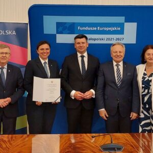 5 elegancko ubranych osób pozuje do zdjęcia. Jedna z kobiet trzyma w ręku otwartą teczkę z jakimś dokumentem. W tle niebieska ścianka z napisem Fundusze Europejskie.