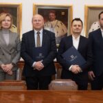 Podpisanie umowy pomiędzy Politechniką a Aeroklubem Krakowskim. 6 elegancko ubranych osób - pięciu mężczyzn i jedna kobieta pozują do zdjęcia na tle ściany z portretami rektorów.