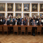 Grupa osób w różnym wieku i różnej płci pozuje do zdjęcia. Część osób siedzi za długim stołem, część osób stoi za nimi na tle ściany z portretami.