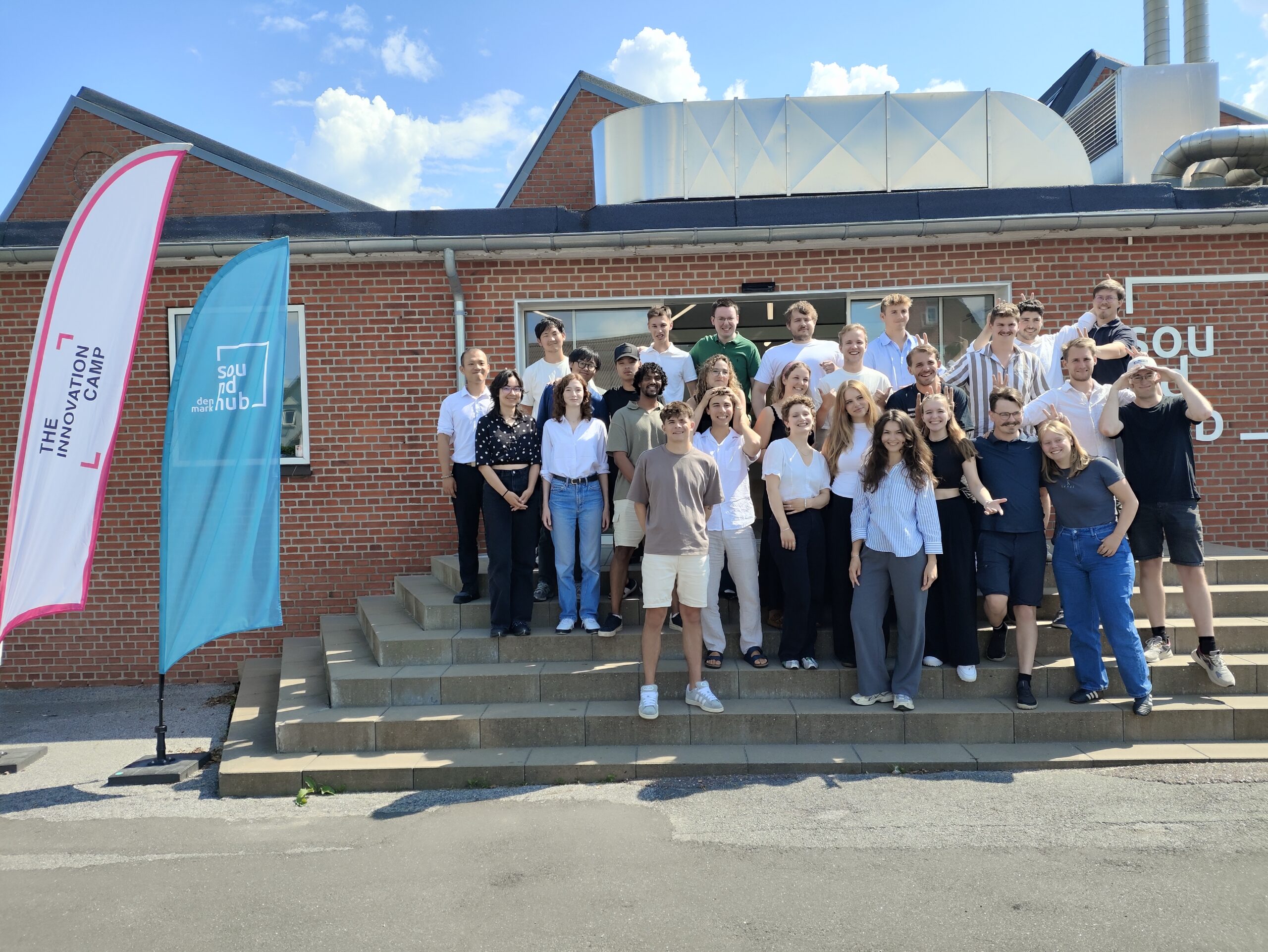 Grupa młodych, uśmiechniętych osób pozuje do zdjęcia na tle niskiego budynku z czerwonej cegły. Z boku 2 windery, na jednym z nich widać napis The Innovation Camp